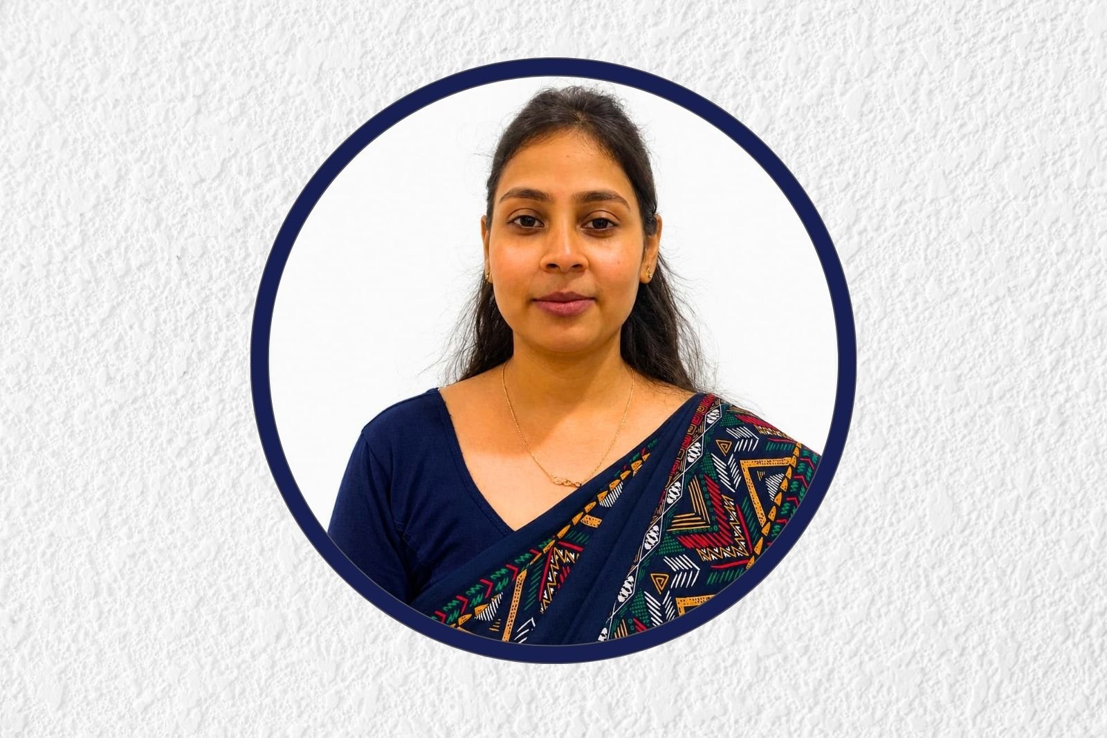 Portrait of a woman wearing a blue sari with a colorful, geometric border, inside a circular navy frame on a white textured background.