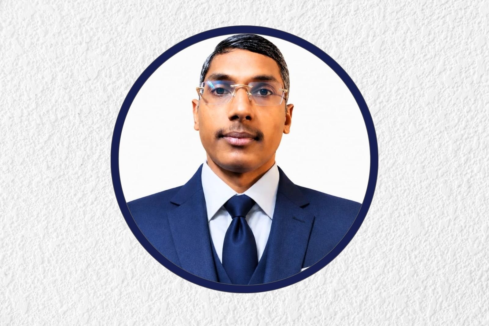 Portrait of a man in a navy suit and tie, wearing glasses, inside a dark blue circular frame on a light textured background.