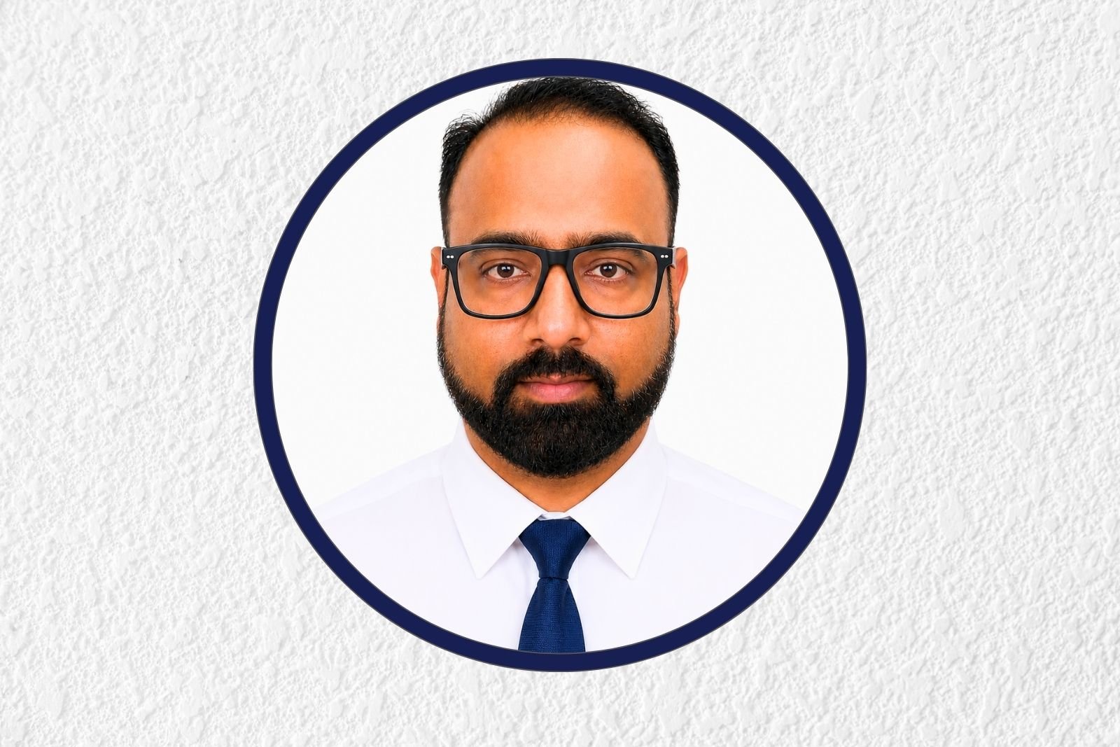 Circular-framed headshot of a man with glasses and a beard, wearing a white shirt and blue tie against a light textured background.