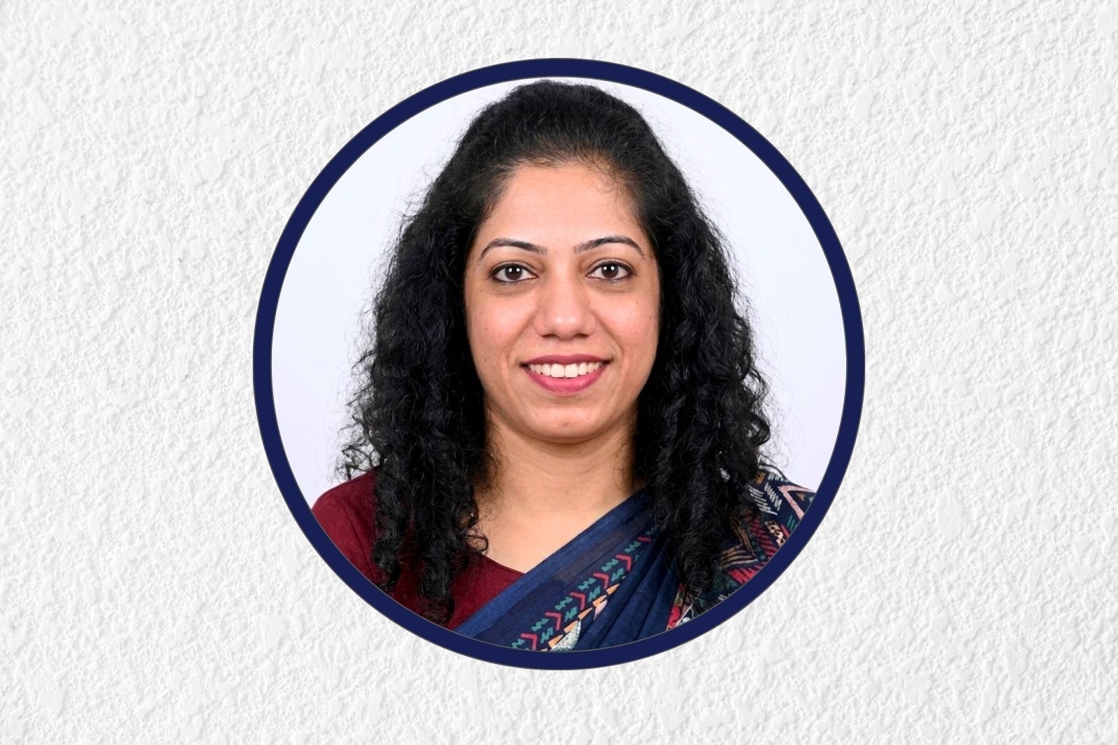 Portrait of a smiling woman with dark, curly hair wearing a blue sari, framed by a navy circular border on a light background.
