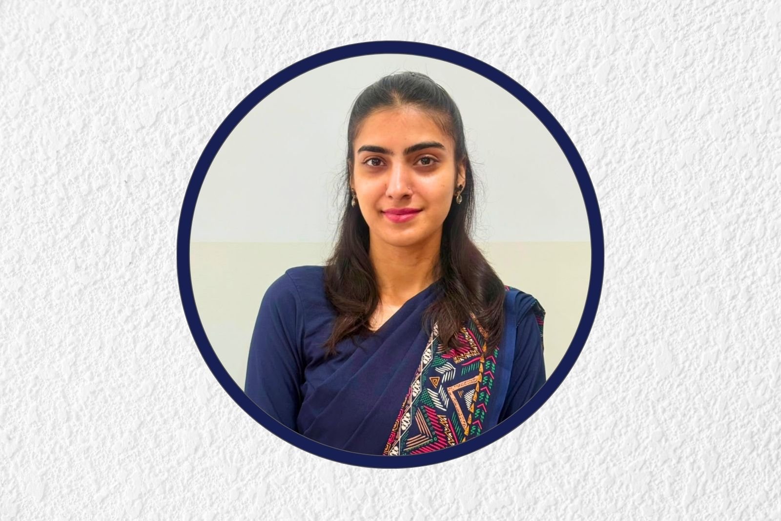 Portrait of a young woman with dark hair in a blue saree and geometric-pattern blouse, centered inside a navy circular frame on a textured light-grey background.