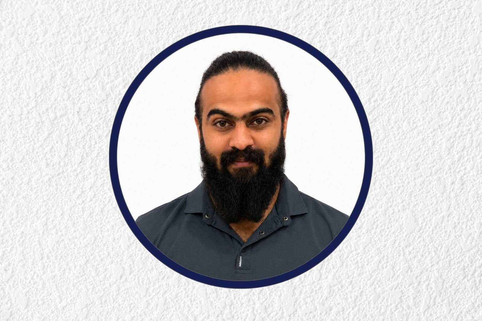 Portrait of a man with a thick dark beard and mustache, wearing a dark polo, inside a circular white frame with a navy border on a textured gray background.