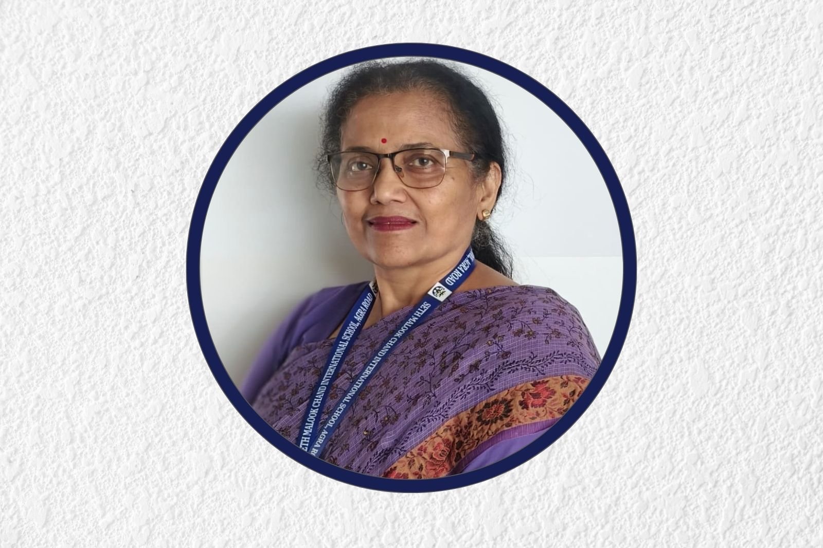 Portrait of a woman wearing glasses and a purple patterned saree with a blue lanyard, framed in a dark blue circle on a textured background.