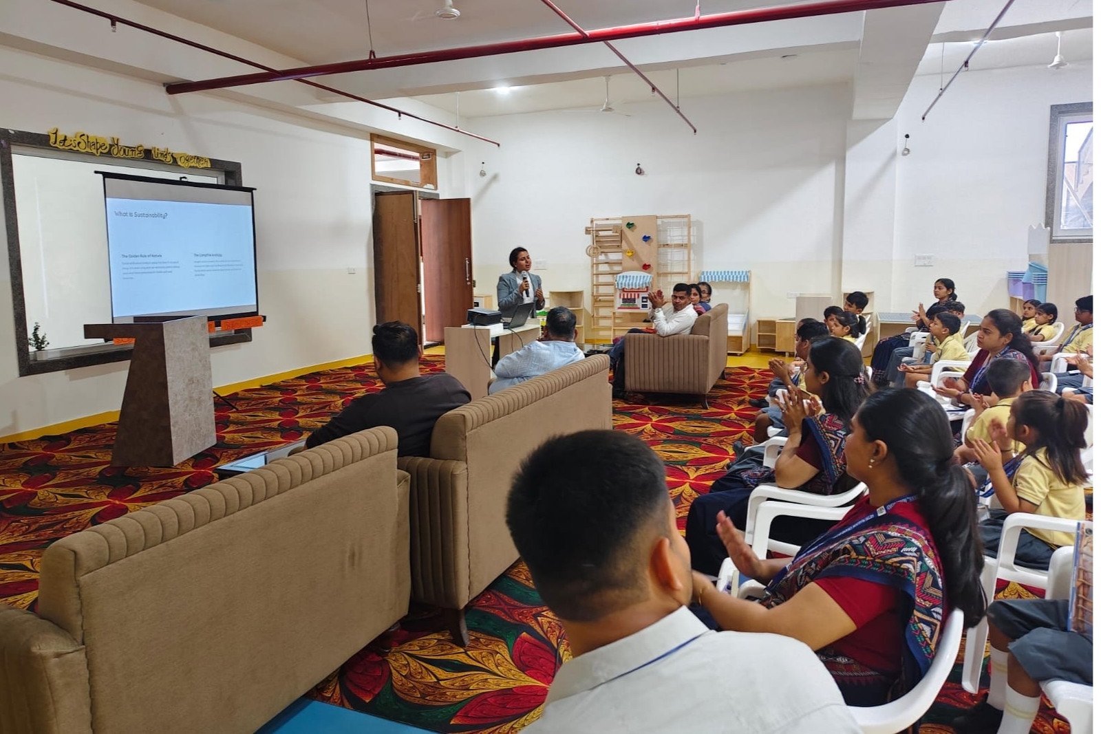 Speaker presenting a slide to a classroom audience of children and adults in a bright seminar room brought together by a facilitator at the front.
